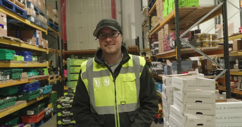 A man with a green reflective vest is standing looking forward and when he is giving a smile there are several shelves behind him