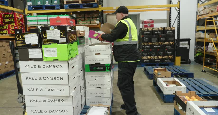 A warehouse worker wearing a high visibility vest and gloves is carefully arranging boxes of produce on a pallet ensuring proper stacking and organization