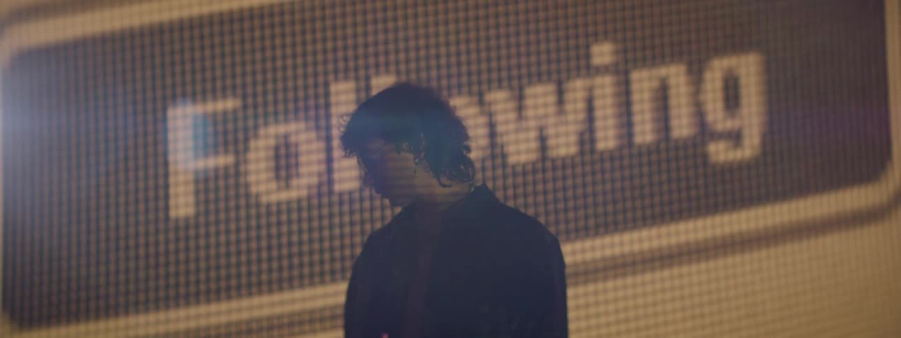A man standing is looking to his right side while behind him there is a huge orange screen with a video of a button being clicked