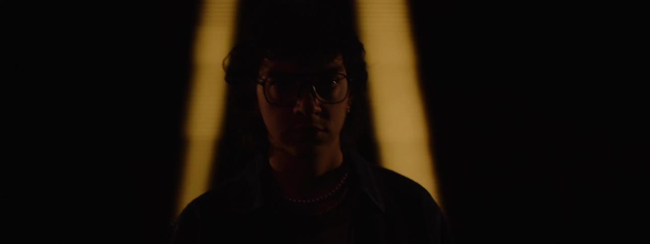 A man wearing glasses and necklaces looks directly at the camera as dim shifting colored lights illuminate their face against a background of abstract light patterns