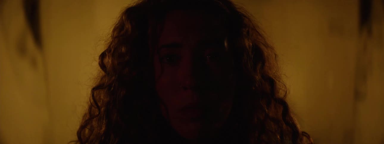 A woman with curly hair stands in front of a screen displaying various faces of people
