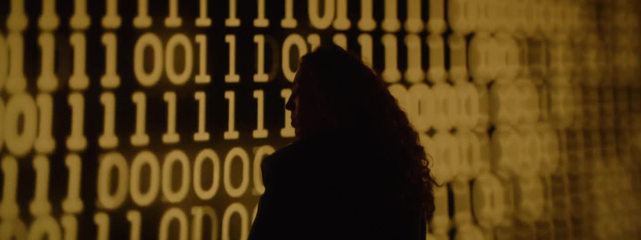 A person stands silhouetted against a glowing wall of binary code creating a futuristic and contemplative atmosphere