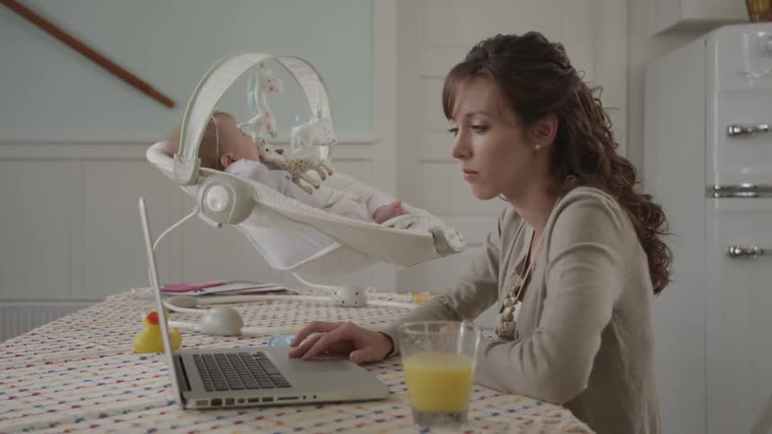 A woman types on a laptop with a baby in a bouncer on the table next to her