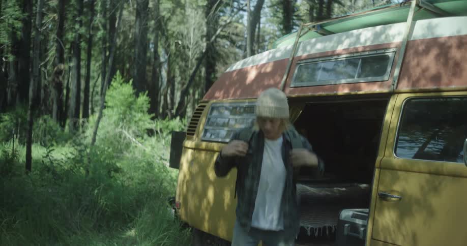 A man wearing a beanie takes a large backpack from his yellow vintage camper van parked in a sunny forest puts it on and walks away