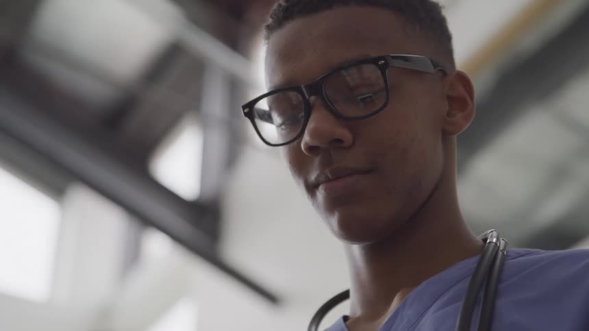 A young black male doctor with a stethoscope hanging around his neck wearing glasses looks down while inside the hospital premises