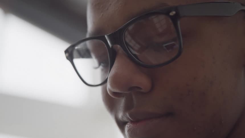 A closeup of a man with glasses his face angled downward inside a welllit room with bright windows
