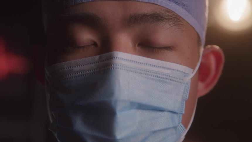 A surgeon wearing a cap and mask slowly lifts his head