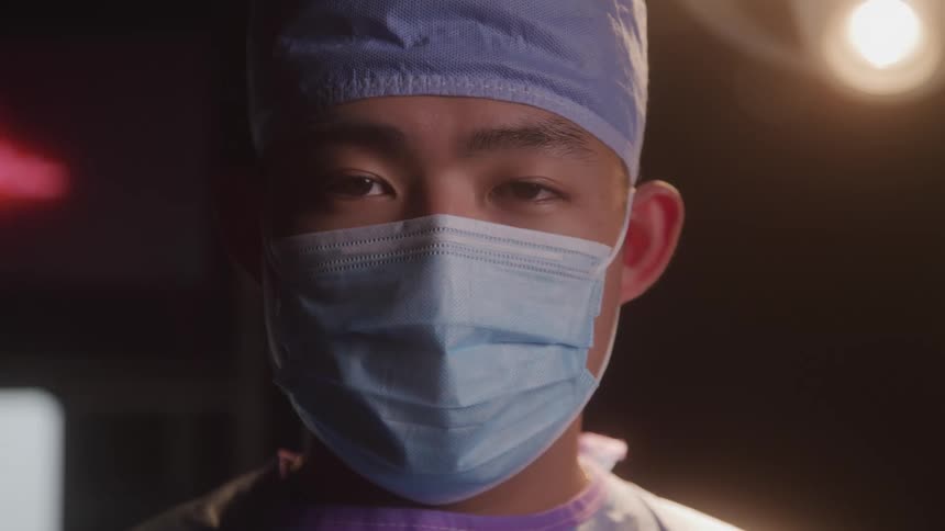 An asian man dressed as a doctor wearing surgical a mask standing in a dimly lit operating room with a heart monitor displaying a heartbeat in the background