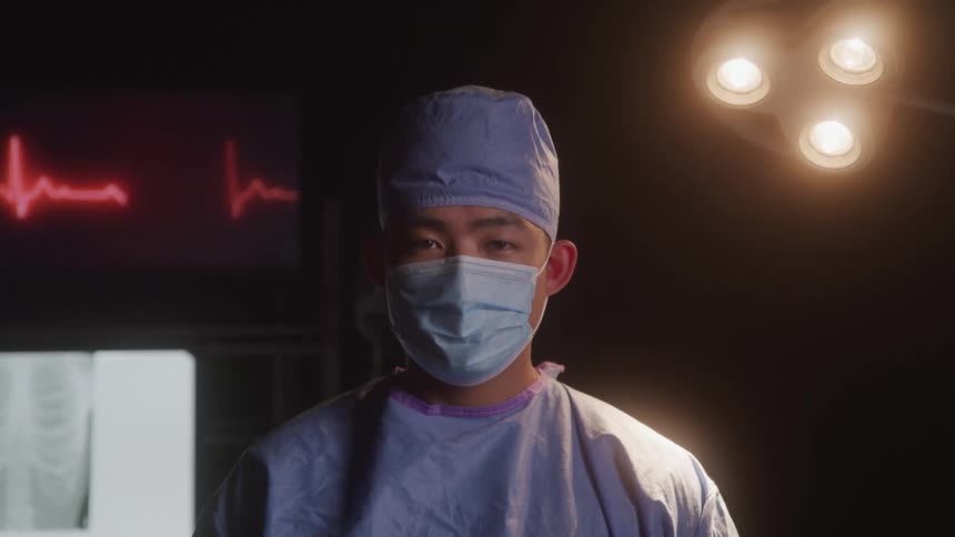 An asian man dressed as a doctor wearing surgical scrubs and a mask standing in a dimly lit operating room with a heart monitor displaying a heartbeat and an xray of a chest in the background