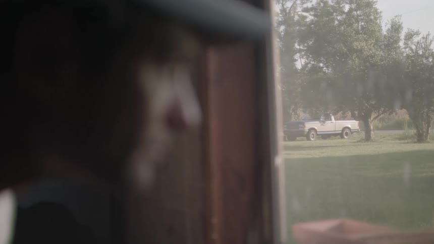 A man wearing a baseball hat looking out a window with a pickup truck parked in the yard on a morning