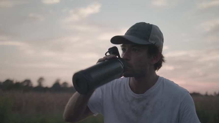 Man in a cap in a field drinking from a metal bottle