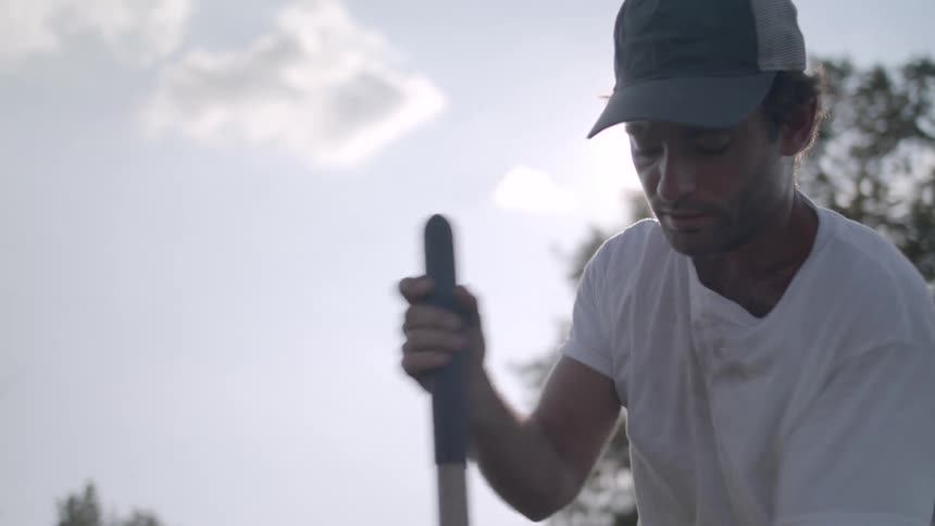 A man wearing a hat and a white tshirt uses a shovel to dig the soil on a sunny day