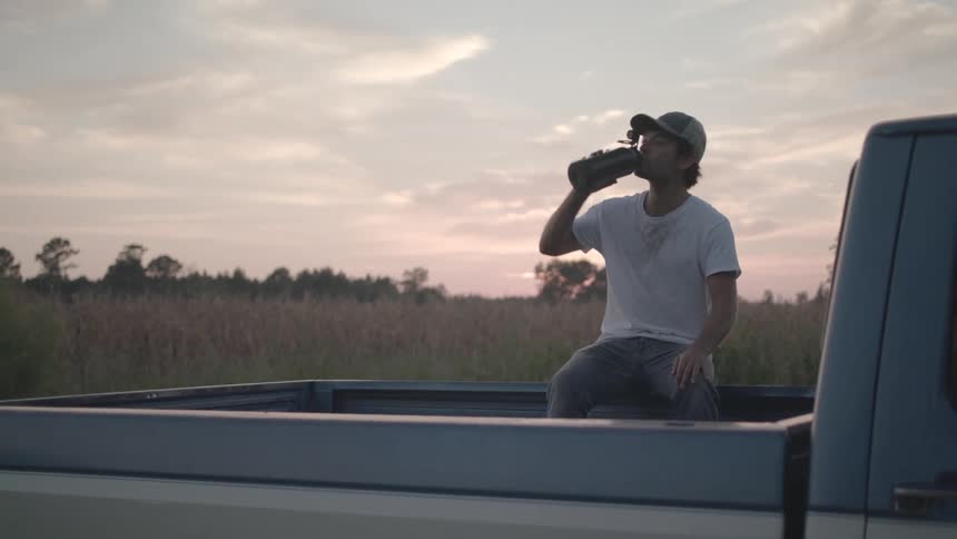A man wearing a white tshirt hat and jeans sits at the back of a truck parked on a grassy field he picks up a water bottle from the truck bed drinks from it and then sets it down at sundown