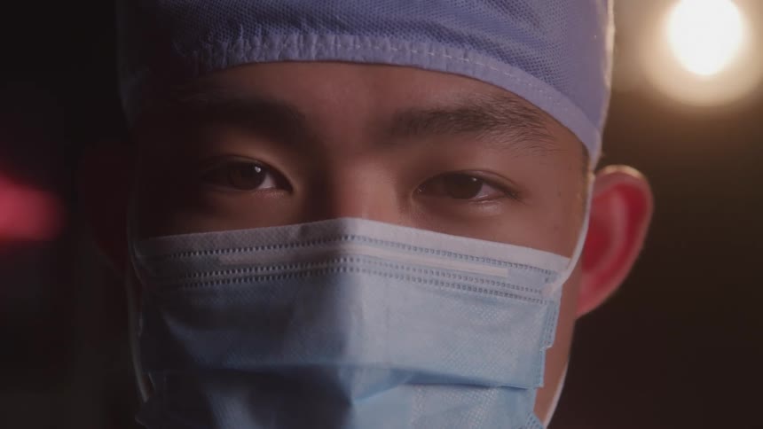 A doctor wearing a nose mask and surgical cap stands with a bright surgical light illuminating the area behind him