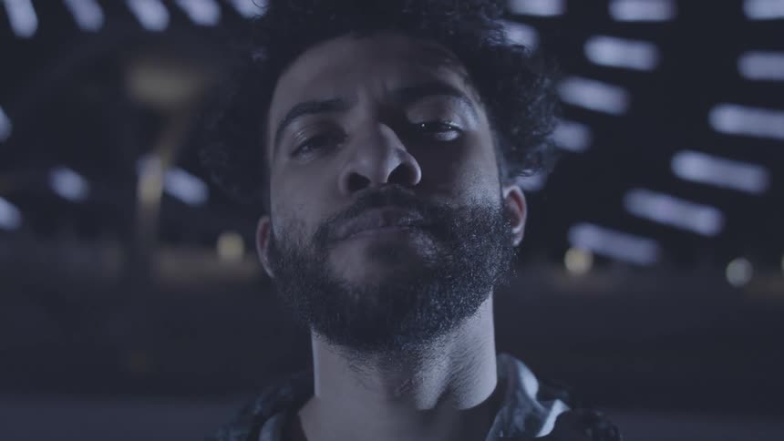 A man with curly hair and a beard slowly looks up at the camera in a dimly lit setting with blurred background lights