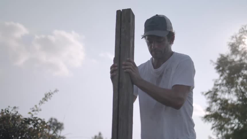 A man wearing a hat and a white tshirt is in a grassland at midday pushing a piece of wood into the soil