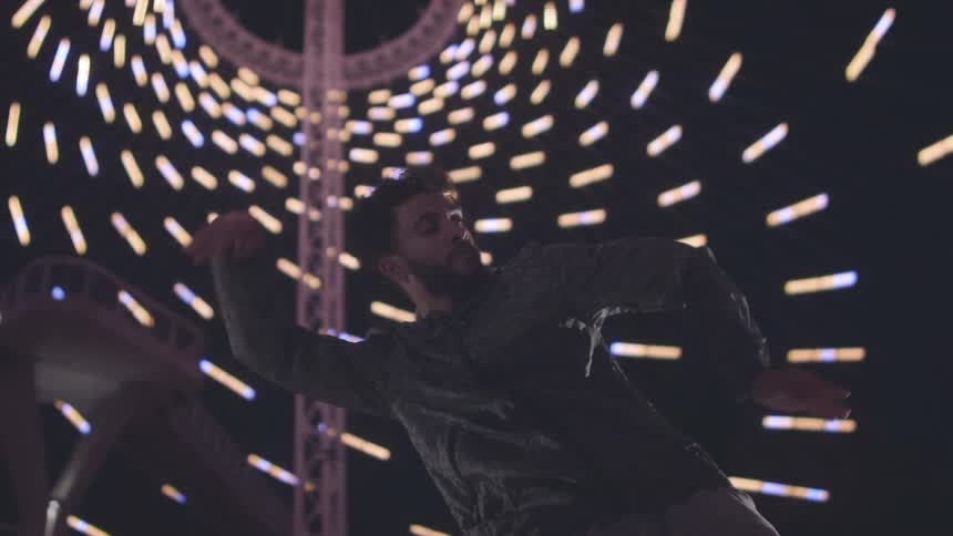 A black man is dancing on stage moving his body and arms rhythmically under a series of lights