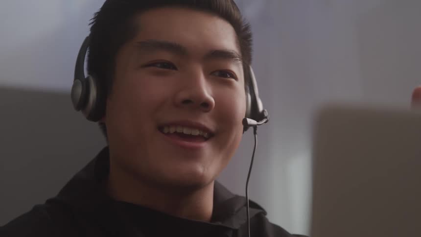 A young adult with dark hair wears a headset with a microphone gesturing animatedly while explaining something in front of a laptop screen