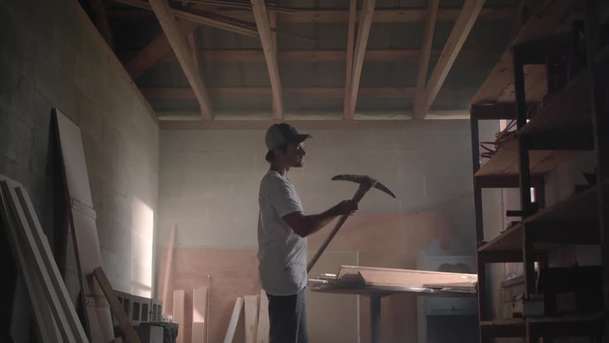 Rewritten a man inside a workshop wearing a hat and a white tshirt picks up a pickaxe to examine it at midday