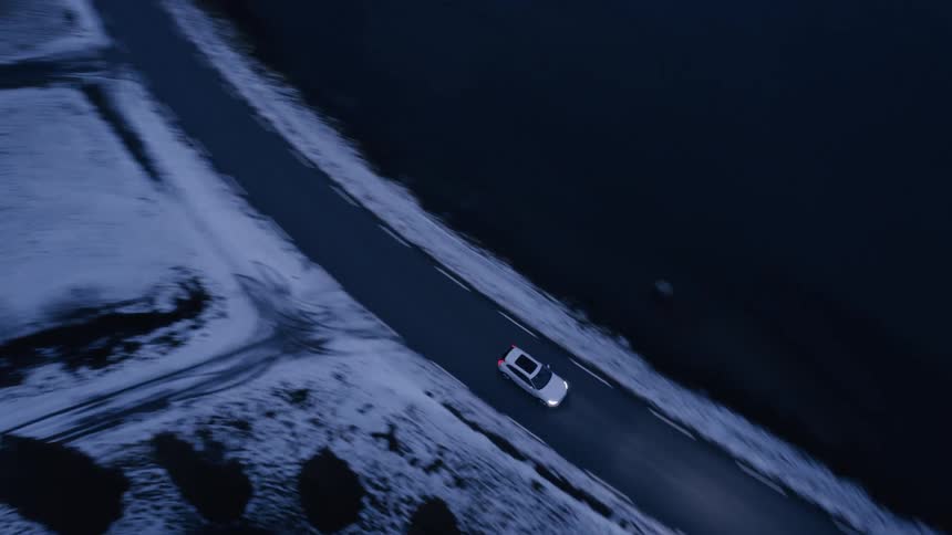 Aerial view of a vehicle traveling along a seaside highway and the edge of snowy mountains