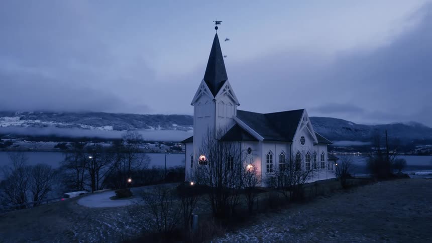 A white wooden church with a dark steeple stands illuminated on a slight hill overlooking a body of water and snowdusted land under a twilight sky