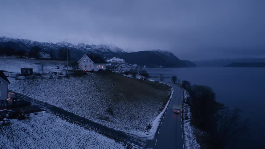 A car on a winter road with a house above the road and snowcovered mountains in the distance