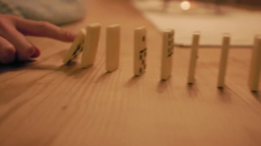 A finger pushes over a line of dominoes on a wooden surface initiating a chain reaction