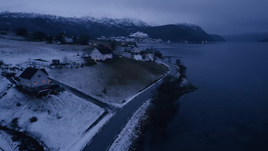 Illuminated houses dot a snowcovered landscape bordering a dark expansive fjord under a dim overcast sky