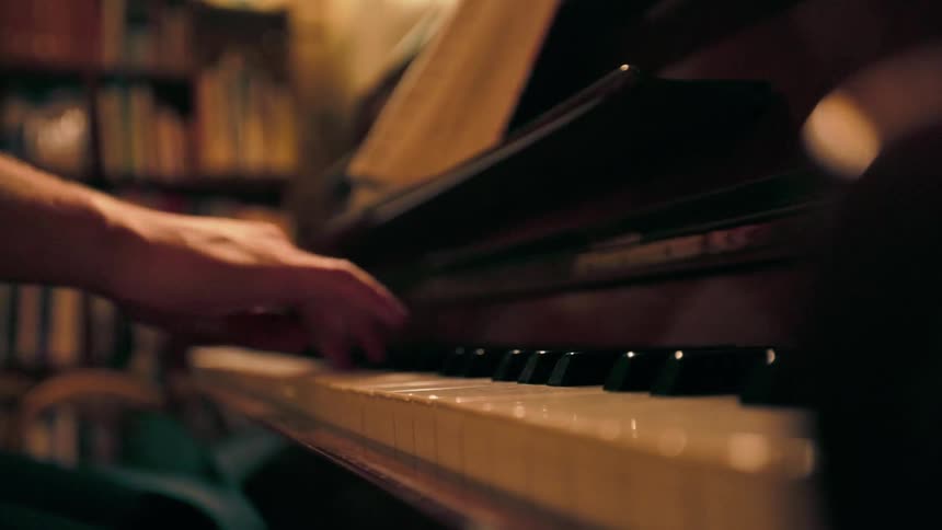 A persons right arm plays a piano with a bookshelf visible on the left side of the room
