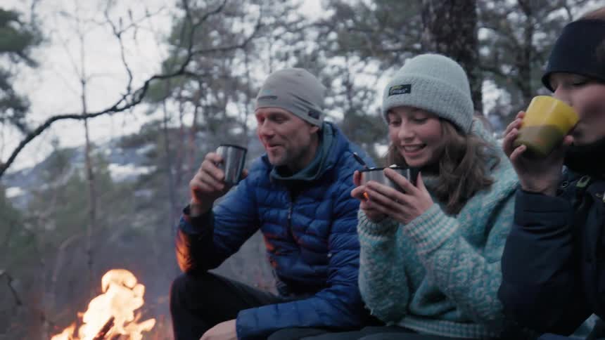A family sits outside near a fire and sips something warm out of a mug