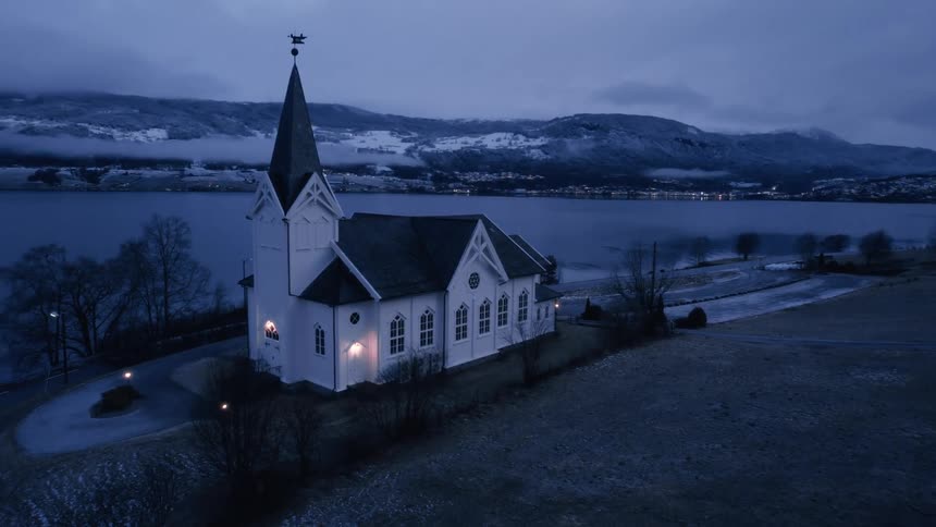 A white steeple church with softly lit windows stands on a snowdusted shore beside a still dark fjord under a cloudy evening sky