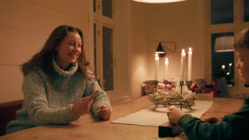 A woman with long brown hair wearing a lightcolored turtleneck sweater smiles at a child sitting across a wooden table adorned with lit candles and playing with dominoes