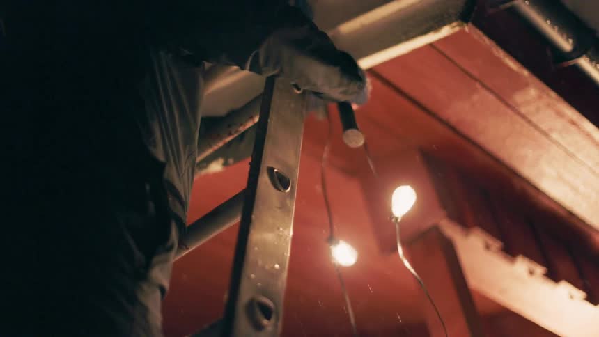 A person on a ladder at the side of a house hangs christmas lights outdoors at night