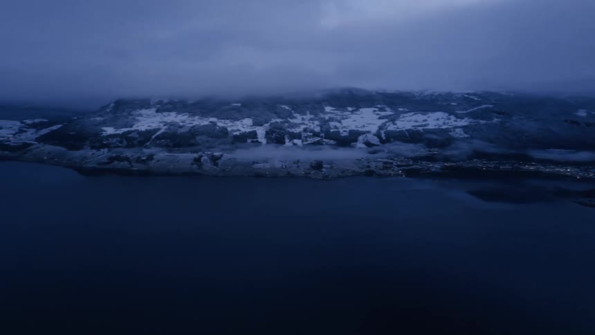 An aerial view at dusk shows a dark lake with distant town lights nestled at the base of snowcovered fogshrouded mountains under a cloudy sky