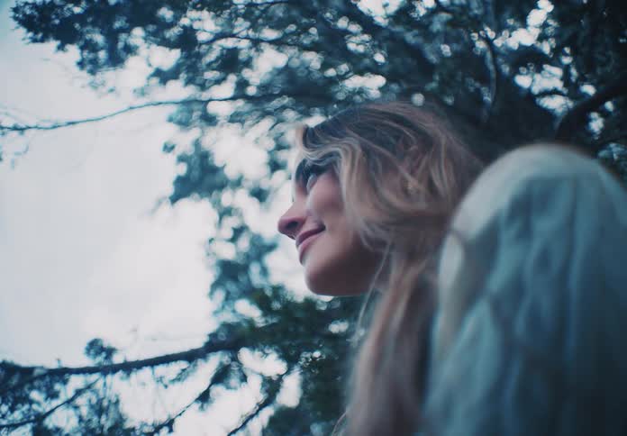 A woman with long lightcolored hair looks smiles amidst the branches of a tree against a muted sky