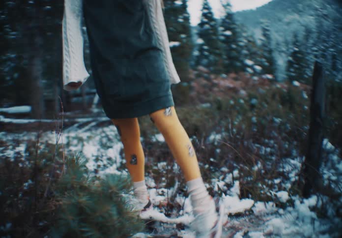 A girl wearing patterned yellow socks and athletic shoes walks on a snowdusted path in a wooded mountainous landscape