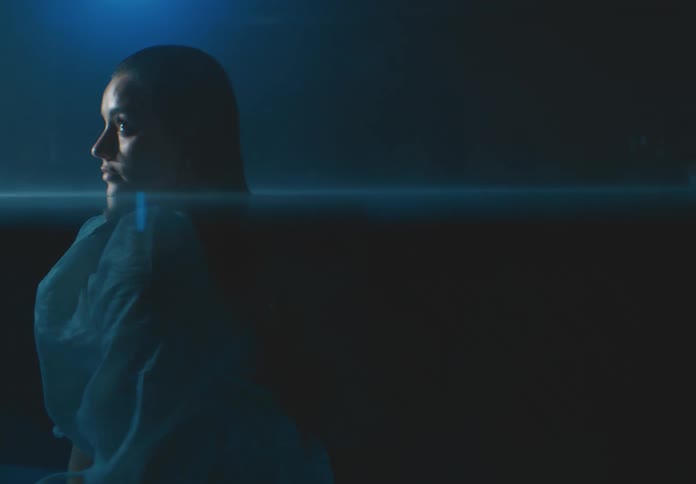 A woman in a white dress floats submerged underwater her head breaking the surface illuminated by a soft dim blue light that envelops the scene