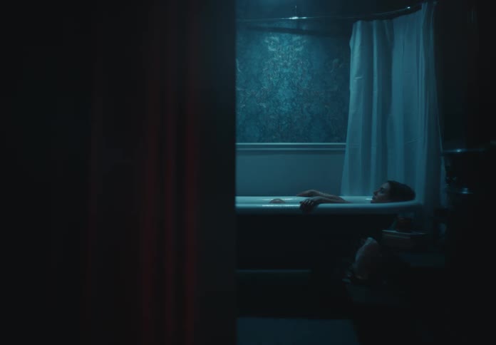 A person is lying in a bathtub in a dimly lit bathroom with patterned wallpaper and a shower curtain