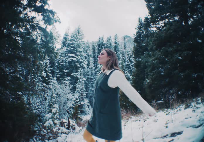 A young woman who is wearing a blue jumpsuit and is walking outside in a snow covered pine forest and she is happy