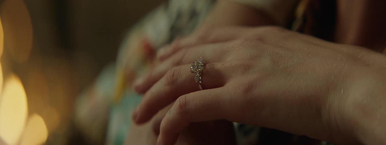 A closeup shot captures a hand adorned with a silver ring featuring a large central diamond surrounded by smaller stones with flickering candlelight casting a warm glow in the softly blurred background