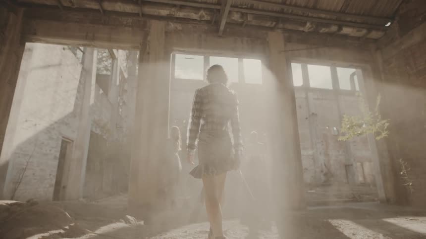 A woman walking like on a runway in an abandoned building under the sunlight while another woman stands nearby leaning against a pillar