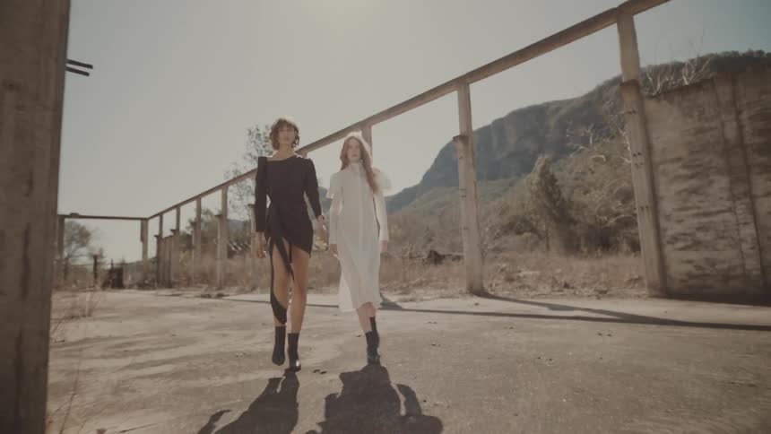 Two women one in a short black dress and one in a long white dress walk through a barebones structure