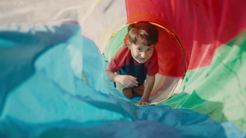 A young boy is crawling through a colorful play tunnel while smiling at the camera