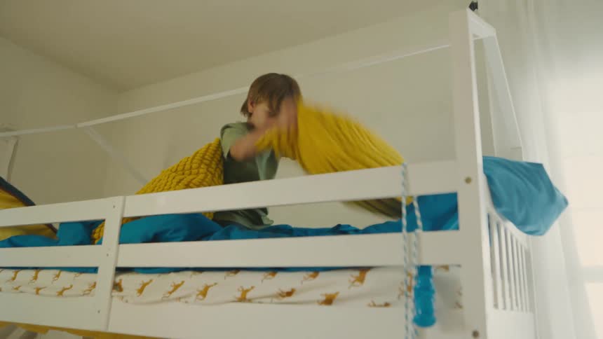 A young boy energetically throws a yellow cushion while playing on the top bunk of a bed