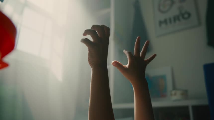 A childs hands reach and gesture playfully toward the light in a softly lit room