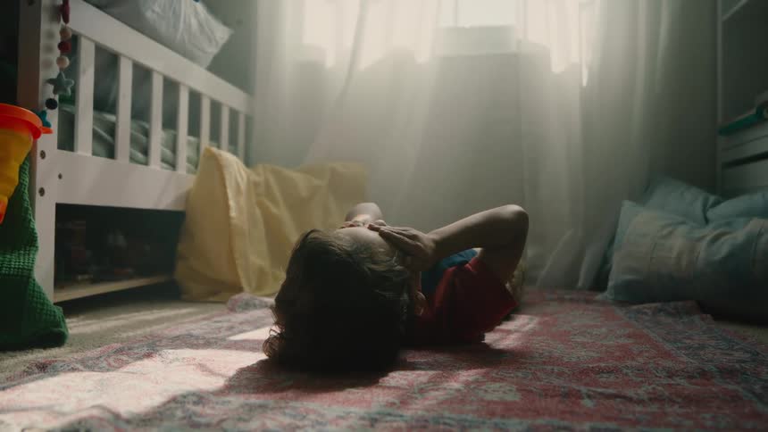 A child lies on their back on a patterned rug in a brightly lit bedroom