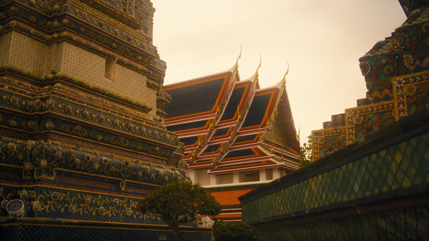 An ornate thai temple complex is captured with a focus on its intricate details and tiered rooftops against a muted sky