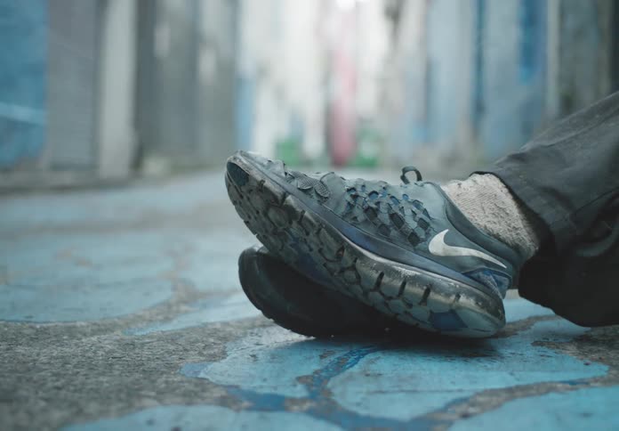 A person wearing worn nike sneakers and dark pants sits on a blue painted street in an alleyway