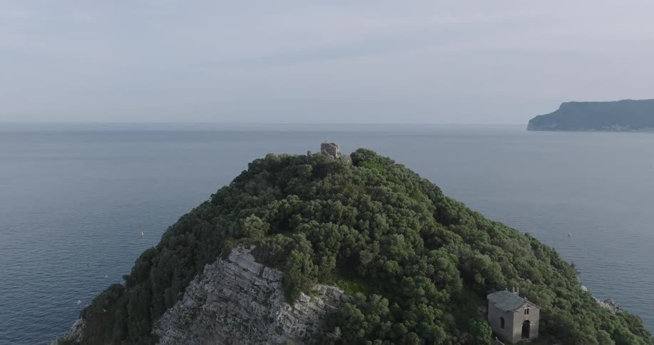 An aerial view of a small lush island with dense trees and the ruins of an old stone structure surrounded by calm expansive ocean waters