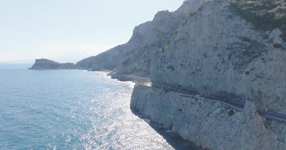 Cars drive along a coastal road carved into the side of a mountain at midday